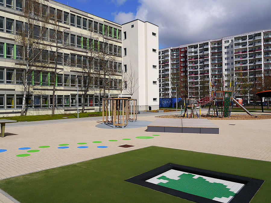 Schulgebäude, über Eck Plattenbau, Hof mit farbigen Punkten auf dem Pflaster und Trampolin