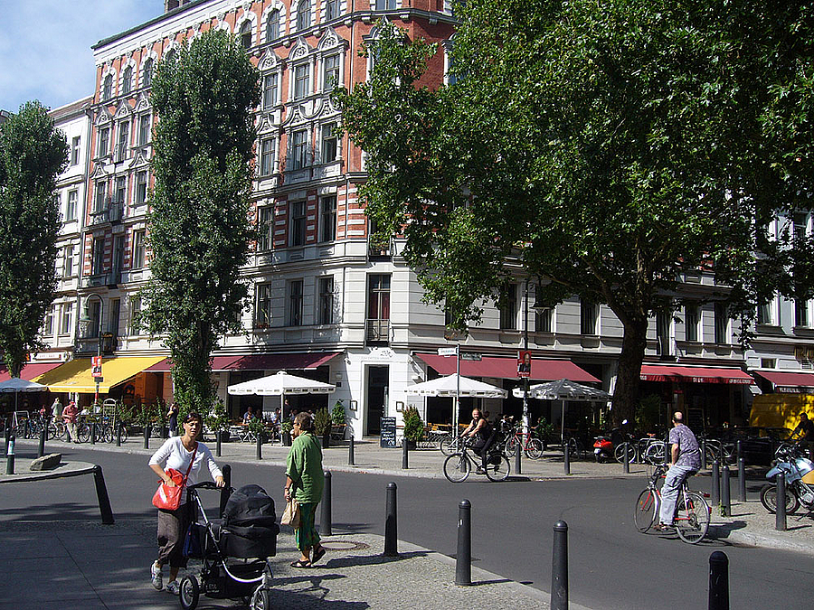 Abgepollerte Straßenkreuzung mit Cafeterrasse, Sonnenschirmen, viele Menschen