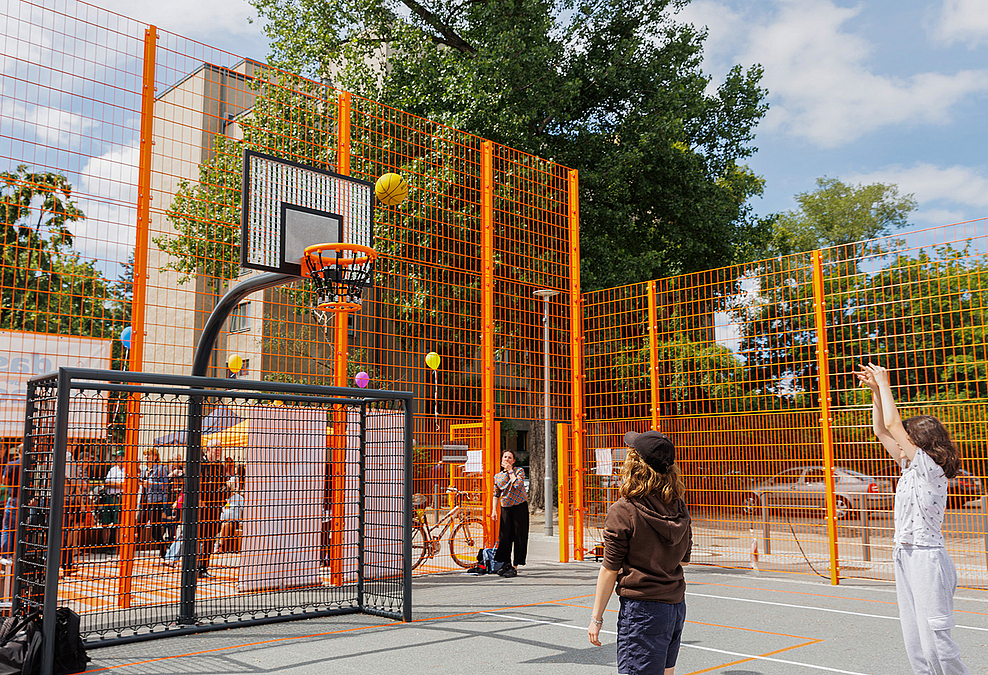 2 Kinder werfen Bälle auf Basketballkorb über Fußballtor, orangefarbener Ballfangzaun