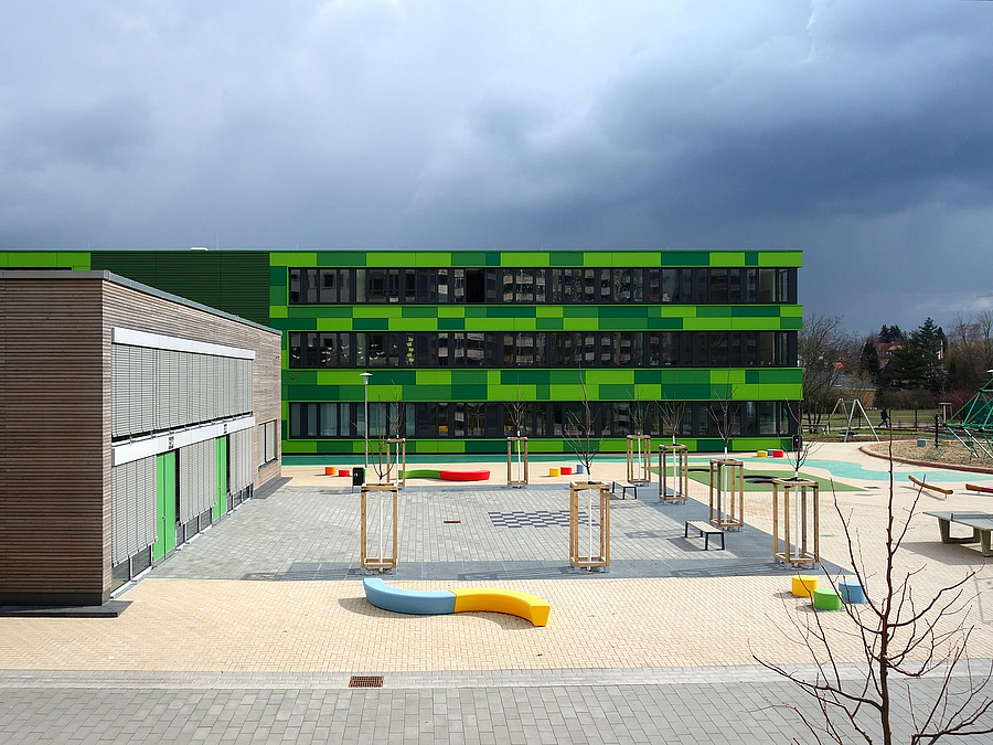 Blick von oben auf Schulhof mit vielen kleineren Elementen, zweigeschossigen Verbinder und grünen Ergänzungsbau