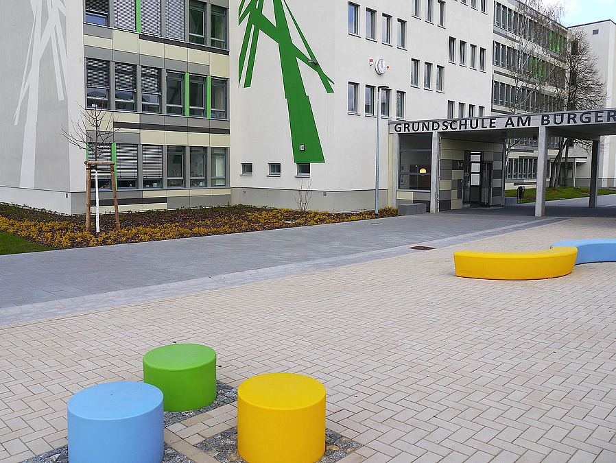 Helles Schulgebäude mit grünem Ornament, Verbindungsgang, gepflasterter Hof, Sitzhocker und -schlange in gelb, grün, hellblau