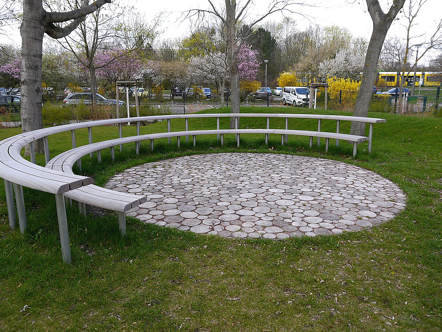 2 stufenförmig angeordnete, halbrunde Sitzbänke um Kreis, gepflastert mit kleinen Baumstammscheiben