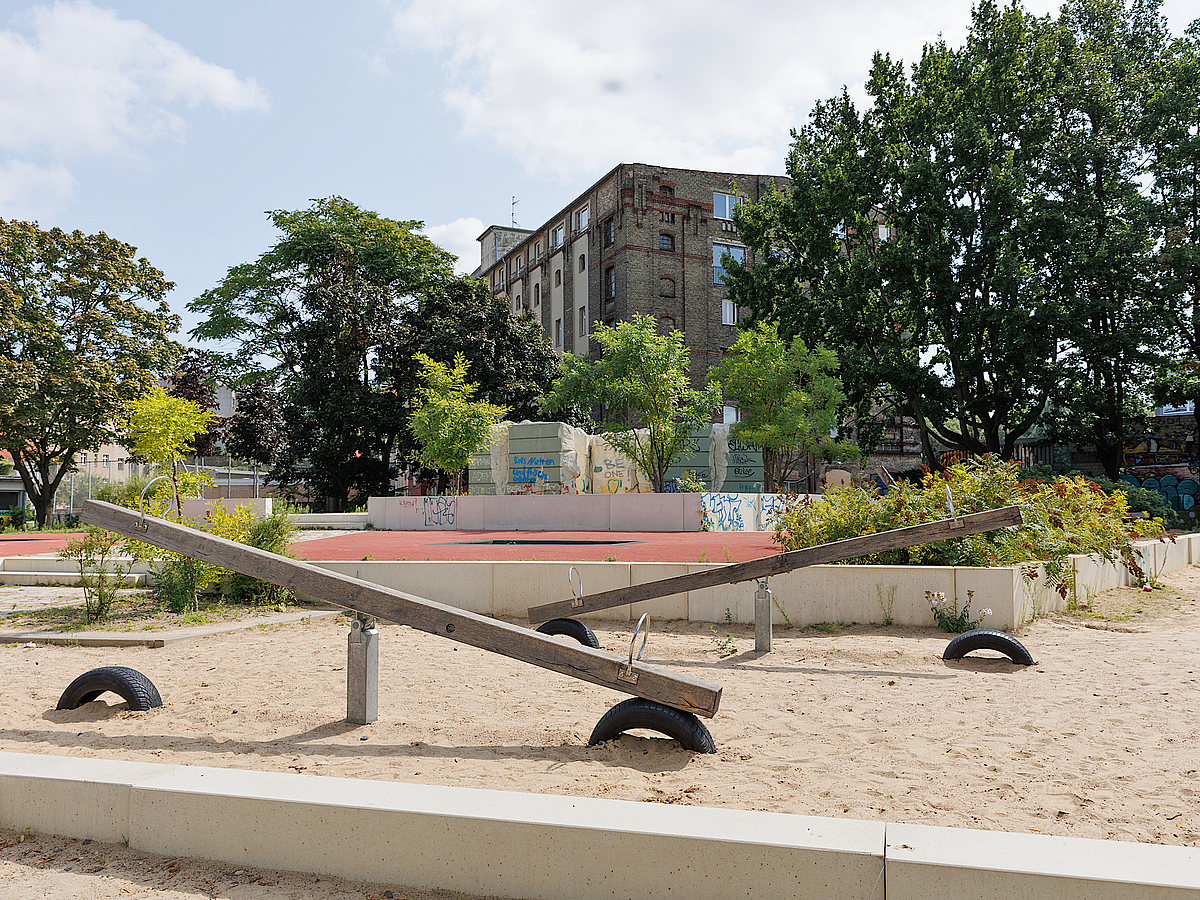 Große Balkenwippen auf Sandspielplatz vor altem Backsteingebäude und großen Bäumen