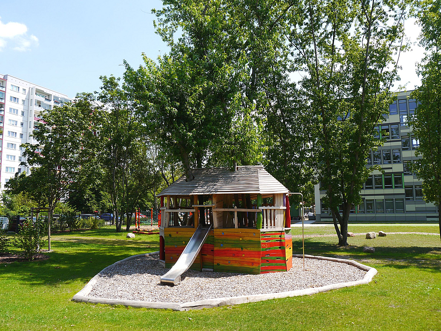 Bunte Hütte auf Holzhäcksel mit Rutsche, Rasen, Bäume. Im Hintergrund modernes Schulgebäude
