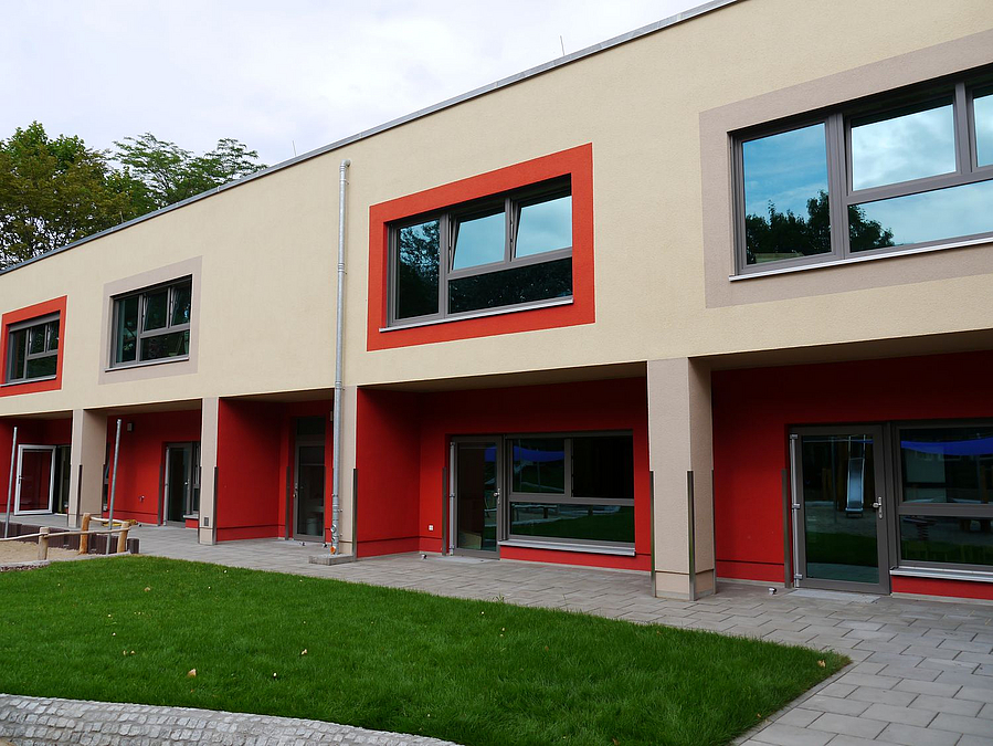 Zweistöckiger beigefarbener Bau mit rot umrahmten Fenstern und Loggien im Erdgeschoss