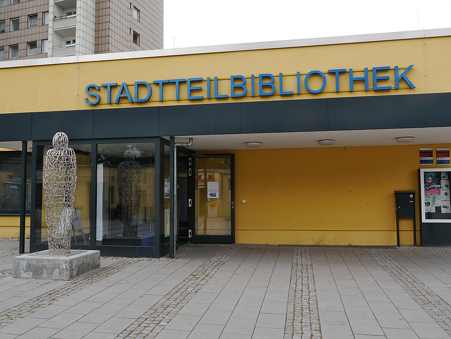 Gelber Flachbau vor Wohnhochhaus mit Schriftzug Stadtteilbibliothek, davor Skulptur aus Draht, eine Person darstellend