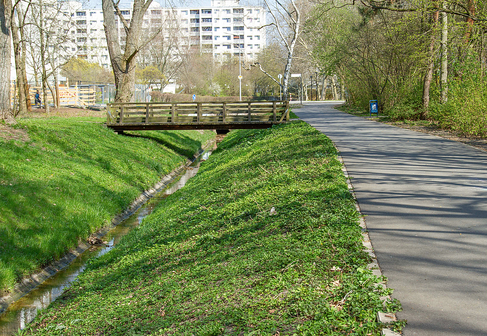 Asphaltierter Weg an Graben mit Brücke, im Hintergrund Hochhäuser