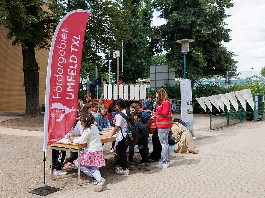 Kinder malen im freien am Stand mit Fahne "Fördergebiet Umfeld TXL"