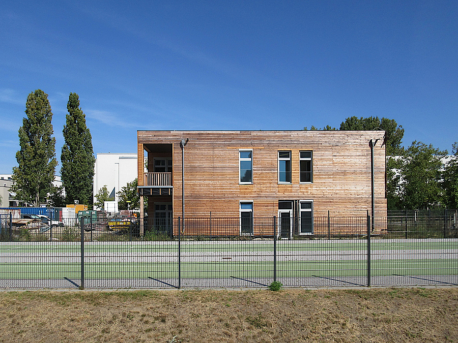 Zweistöckiger Holzbau hinter Zaun und grüner Laufbahn