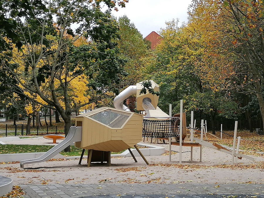 Großes Spielgerät in Beige mit vieleckigen Elementen, Rutschen und Kletternetzen, große Bäume