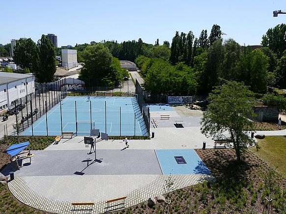 Blick auf befestigten Platz mit Streetballmast, Bolzplatz mit Ballfangzaun und blauem Boden, umsäumt von Grün 